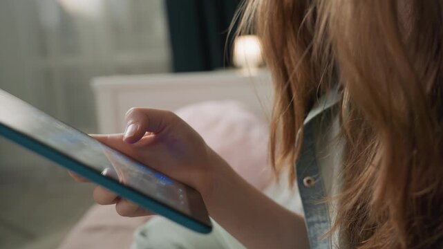 Closeup hands holding tablet young student scrolling apps on blue tablet while sitting on bed in denim shirt, soft lamp glow and pastel pillow in background, relaxed evening mood, fingertip tapping