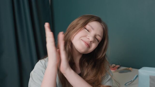 White woman clapping hands smiling at desk, eyes closed in contentment, long brown hair, relaxed cardigan outfit, teal wall and curtain backdrop, bedside table with beauty box and accessories, playful