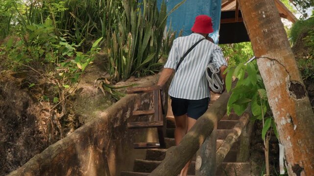 Man carrying wooden crate up stairs, reddressed woman leads, rustic stone steps with handrail, blue tarp shelter and tropical plants, porter movement suggests tea ceremony or vendor delivery, focused