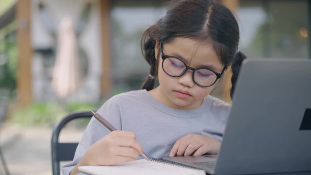 Young girl with glasses writing in notebook outdoors, focused on schoolwork, surrounded by nature, sunny day, learning, inspiring future generations of scholars