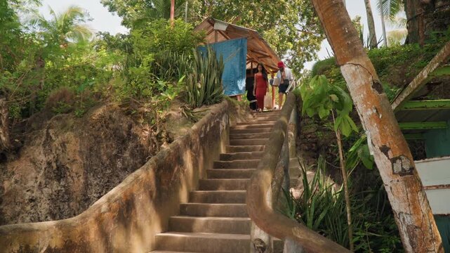Couple descending coastal steps carrying tea, woman in red dress and man in striped shirt with red hat walk down mossy stone staircase toward beach hut amid lush palms, intimate atmosphere, ritual