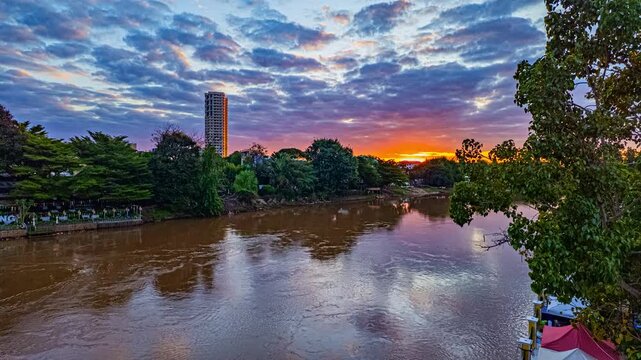 Under a canopy of soft, drifting clouds, at Ping river glistens in the fading golden light of sunset. Gentle ripples reflect shades of orange and pink, while lush greenery hugs the peaceful shoreline