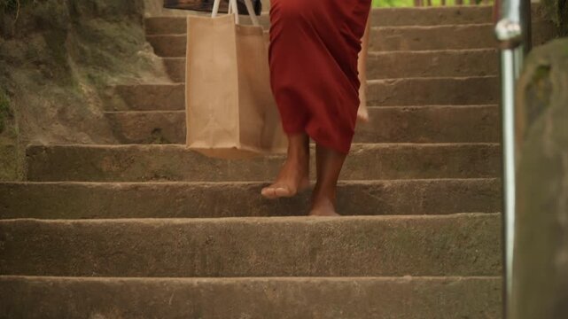 Graceful woman in scarlet dress steps quietly through lush garden. Serene scene of woman in crimson gown walking softly along stone stairs in peaceful garden surroundings with sunlight