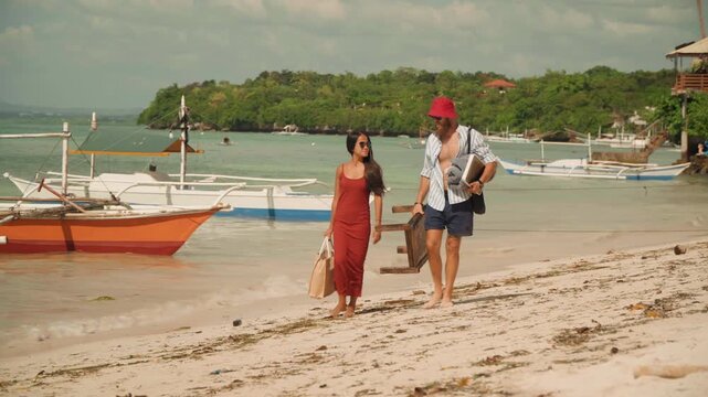 Couple walking on beach holding tea, man in red hat carries wooden tea chest, woman in red dress with straw bag, traditional outrigger boats anchored at shoreline, warm sunlight, relaxed romantic