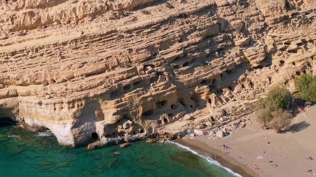 Aerial close-up of Matala beach caves at sunset, Crete. Drone view of tourists walking inside ancient roman caves in sandstone cliff. Scenic golden hour travel destination in Greece, 4k video.