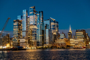 Naklejka premium Hudson Yards Manhattan skyline with crane and night lights. Night cityscape of Hudson Yards district featuring a construction crane and brightly lit glass towers from the water.