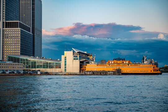 Staten Island Ferry docked at Manhattan terminal under sunset sky. View of the orange ferry at the pier with financial district buildings and colorful clouds in background.