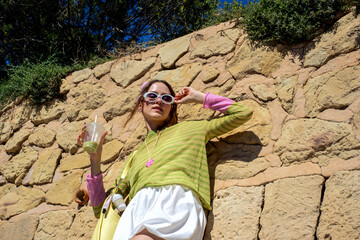Fototapeta premium Young woman enjoying refreshing drink, wearing trendy sunglasses and colorful outfit against stone wall on sunny day