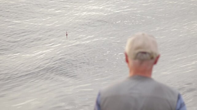 Man in a cap stands by the water's edge, focused on a small float bobbing on the tranquil surface. The scene captures a moment of quiet concentration and connection with nature, evoking a sense of pea