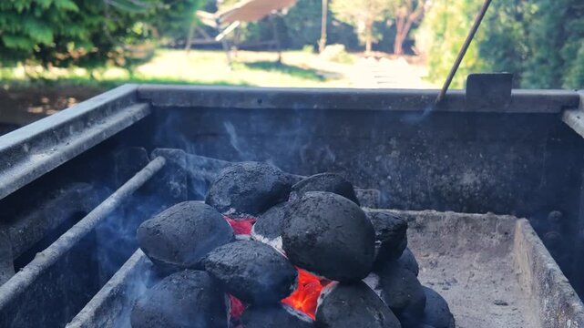 Close up of burning charcoal briquettes in a grill, hot coals with smoke for outdoor barbecue party in a garden.