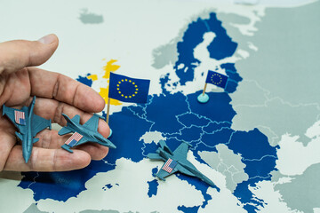 Naklejka premium Hand withdrawing miniature toy fighter jets, representing United States troops, from a European map with European Union flags, symbolizing military withdrawal and changing geopolitics