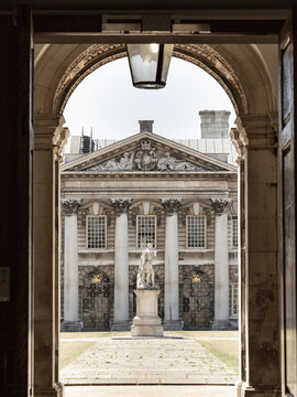 Framed by the dark silhouette of a majestic archway, the image reveals a sun-drenched courtyard that serves as a stage for classical grandeur. A regal marble statue stands atop a weathered stone plint