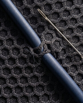 A vertical macro study capturing the interaction between a deep charcoal neoprene honeycomb weave and a smooth matte navy polymer wire, crossed by a precise metal sewing needle.