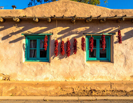 Adobe house wall with traditional turquoise painted window frames and hanging red chili ristras drying in sun.