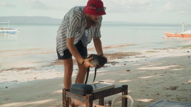 Man preparing tea on beach, setting kettle on low bench, pouring hot water into cup, gentle waves and boats in background, warm morning light, casual striped shirt and red hat, relaxed ritual mood