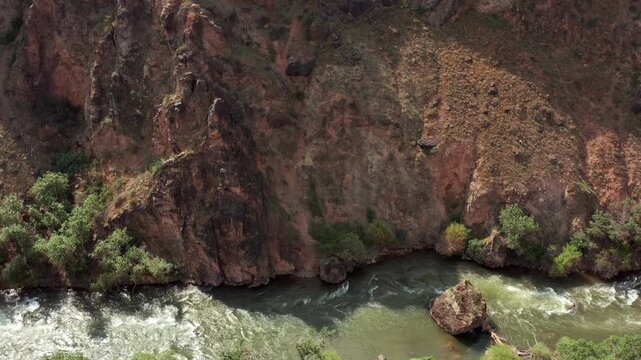 Aerial tilt shot over dramatic dark canyon cliffs with fast flowing river below, strong contrast of rock textures and water, cinematic nature landscape, power, scale and wilderness concept