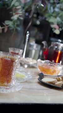 Traditional tea ceremony featuring black tea in armudu glasses, a glass teapot, lemon slices, jam, and a variety of nuts and dried fruits on a marble table. Vertical orientation.