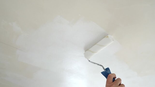 A worker applies white paint to the ceiling during an apartment renovation, painting the surface