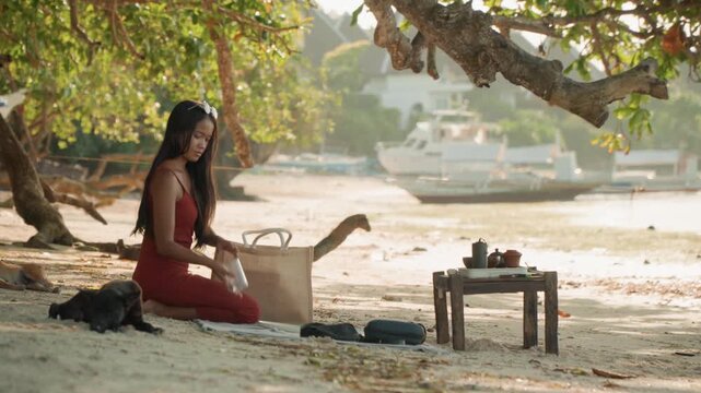 Asian woman on beach preparing tea slow ritual under tree shade with small wooden table teapot cups woven basket soft sunset light gentle waves moored boats in background peaceful meditative mood