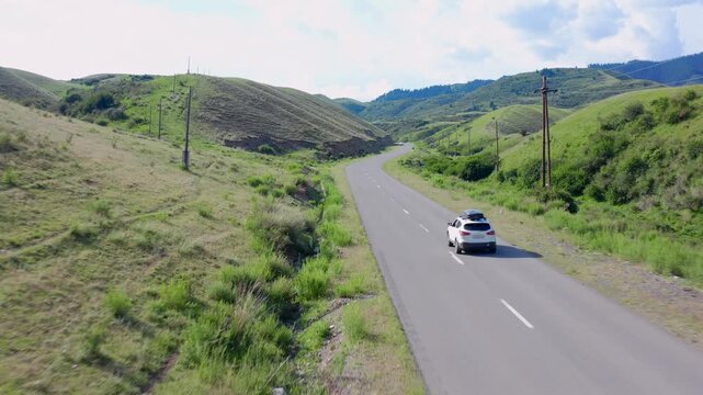 Aerial tracking shot of car driving along winding mountain road near Zhalanash and Saty, surrounded by green hills and Tien Shan landscape in Kazakhstan, scenic rural travel route