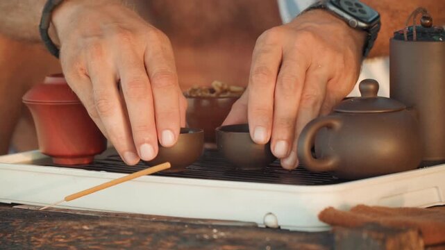 Seaside tea ceremony at sunset closeup hands preparing clay teapot and tiny cups on rustic wooden tray, gentle pour into cup, warm golden light, relaxed beach atmosphere, aromatic herbal infusion,