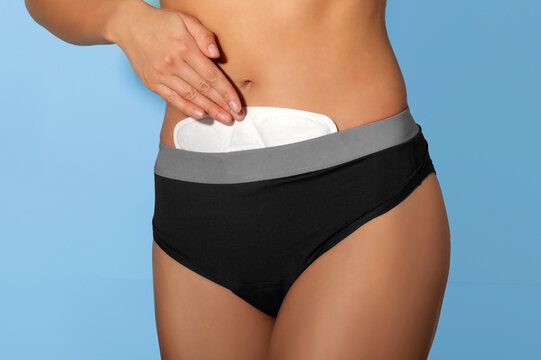 Woman wearing period underwear and applying heating patch to her lower abdomen to relieve menstrual cramps, blue background, closeup. Modern pain relief during menstruation