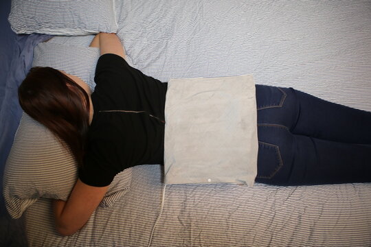 High angle view of a woman lying on a bed using a grey electric heating pad on her lower back for pain relief and relaxation.