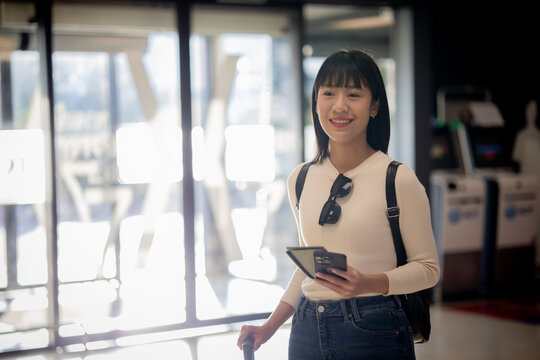 Smiling young Asian traveler holding smartphone and passport at airport terminal
