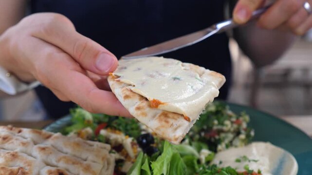 Close up of a person spreading delicious hummus on a warm piece of pita bread with a knife, with a fresh mediterranean salad with feta cheese and tabbouleh in the background