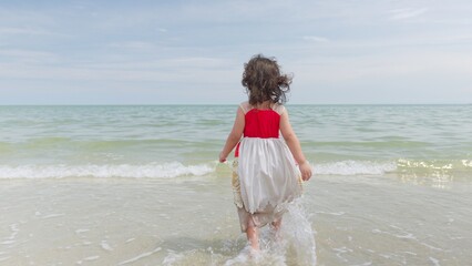 A curious young girl stands in the shallow ocean waves, looking out at the vast horizon, symbolizing childhood wonder and a peaceful moment of discovery. © HarryKiiM Stock