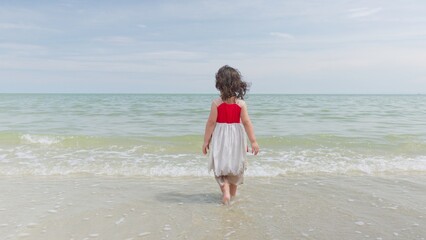 A curious young girl stands in the shallow ocean waves, looking out at the vast horizon, symbolizing childhood wonder and a peaceful moment of discovery. © HarryKiiM Stock