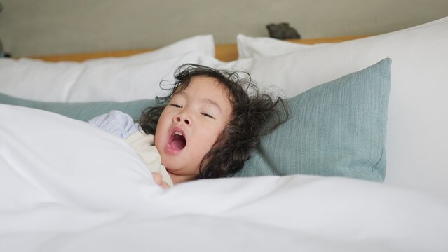 A sleepy young child yawns widely while waking up in a comfortable white bed, a candid moment of childhood fatigue representing rest and domestic wellbeing.