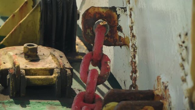 Rusted red chain on deck, closeup of winch and corroded pulley flaking paint, oxidized bolts, weathered metal and industrial textures suggesting deckhand inspection, rigging maintenance, marine