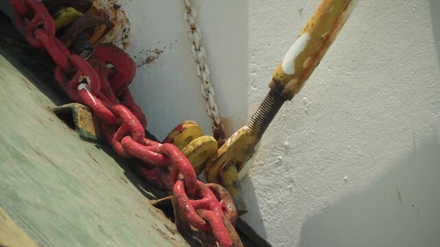 Red chain yellow shackle on deck angled closeup showing mooring point with corroded paint, green planking, heavy link under tension, rugged industrial atmosphere, seaside maintenance context