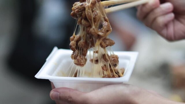 Close up of person using chopsticks to pull sticky fermented natto soybeans from small white container creating stringy texture.