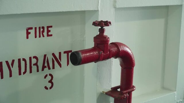 Red fire hydrant mounted on wall closeup of glossy valve and pipe against white painted panel stenciled FIRE HYDRANT industrial safety inspection maintenance mood metal texture bolts slight rust