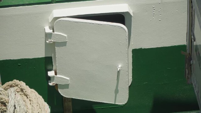 White hatch on green hull, sunlit closeup showing rope coil and metal hinge, chipped paint and salt stains on rough fiberglass surface, mooring detail of fishing vessel at dock, quiet maritime