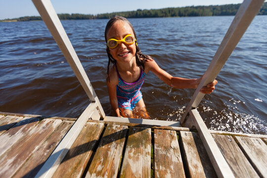Triumphant child emerges from lake through wooden dock completing successful swim on summer day. Little girl surfaces with satisfied expression after accomplished open water experience. Kid feels.