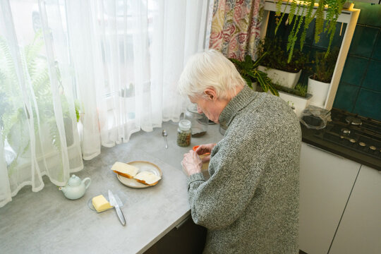 Top-down view of elderly man with stooped back preparing butter sandwiches. Overhead perspective shows arrangement of food and focused activity. Grandpa at kitchen counter.