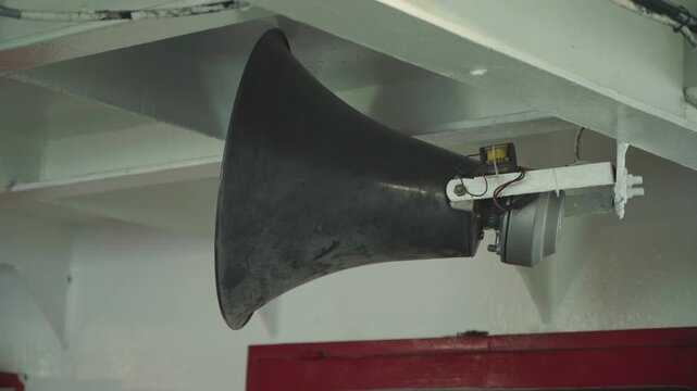 Ship horn mounted under deck beam black conical loudspeaker fixed to white overhead bracket inside cabin red bulkhead and steel mount visible closeup shot capturing corrosion, paint texture, bolts,