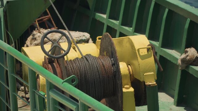 Yellow winch on ship deck, rusted cable spool and wheel handle, green railings framing heavy machinery, maintenance scenario with engineer tools, sunlit weathered paint and metal textures, closeup