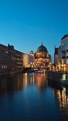 Fototapeta premium Historic buildings line a canal in Berlin during dusk, showcasing illuminated facades and reflections on the water's surface under a clear blue sky