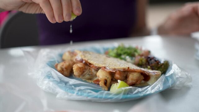 Mature woman eats shrimp tacos from a taco stand in Baja California Mexico. She adds ingredients to enhance the flavor of her meal.