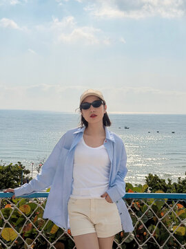 Young woman stands by the sea wearing a striped shirt, shorts, and sunglasses, enjoying a sunny day on a beach with calm waves nearby