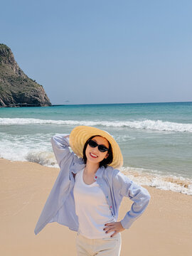 Woman enjoys sunny day on beach while wearing sunglasses and straw hat near water with waves and cliffs in background