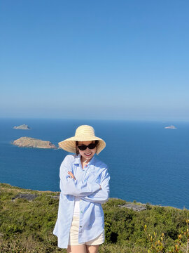Woman stands on a hill with a view of the ocean and islands while wearing a large hat and sunglasses, enjoying a sunny day near the coast