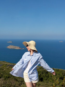 Young woman enjoying the view from a coastal cliff wearing a hat while standing with open arms on a sunny day near the ocean