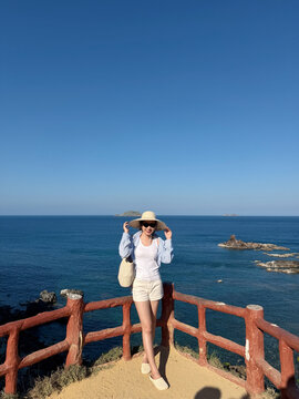 Young woman enjoys a sunny day at the shore while wearing a wide-brimmed hat, standing on a wooden lookout near the ocean during daytime