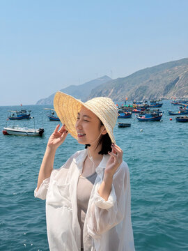 Young woman wearing a straw hat smiles at the seaside with fishing boats in the water on a sunny day near the mountains