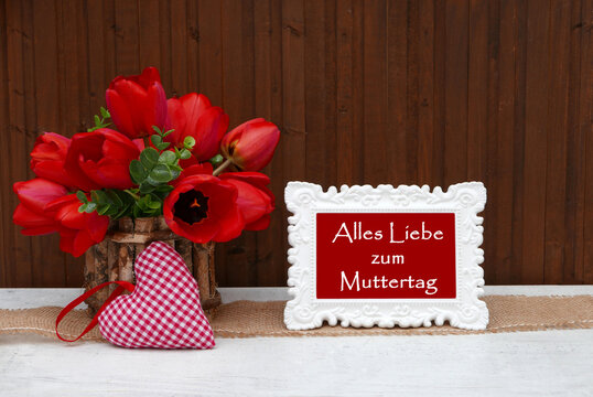 Vase mit roten Tulpen, einem rot kariertem Stoffherz  und Bilderrahmen mit Text 'Alles Liebe zum Muttertag' vor Hilzhintergrund.